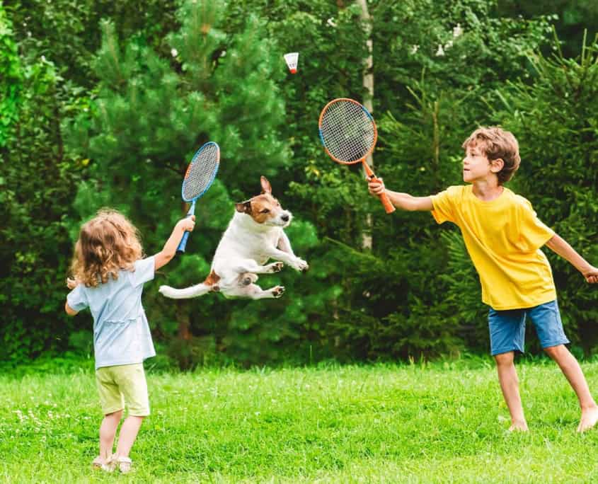 Kinder spielen Federball auf der Wiese und ein Hund springt in die Luft